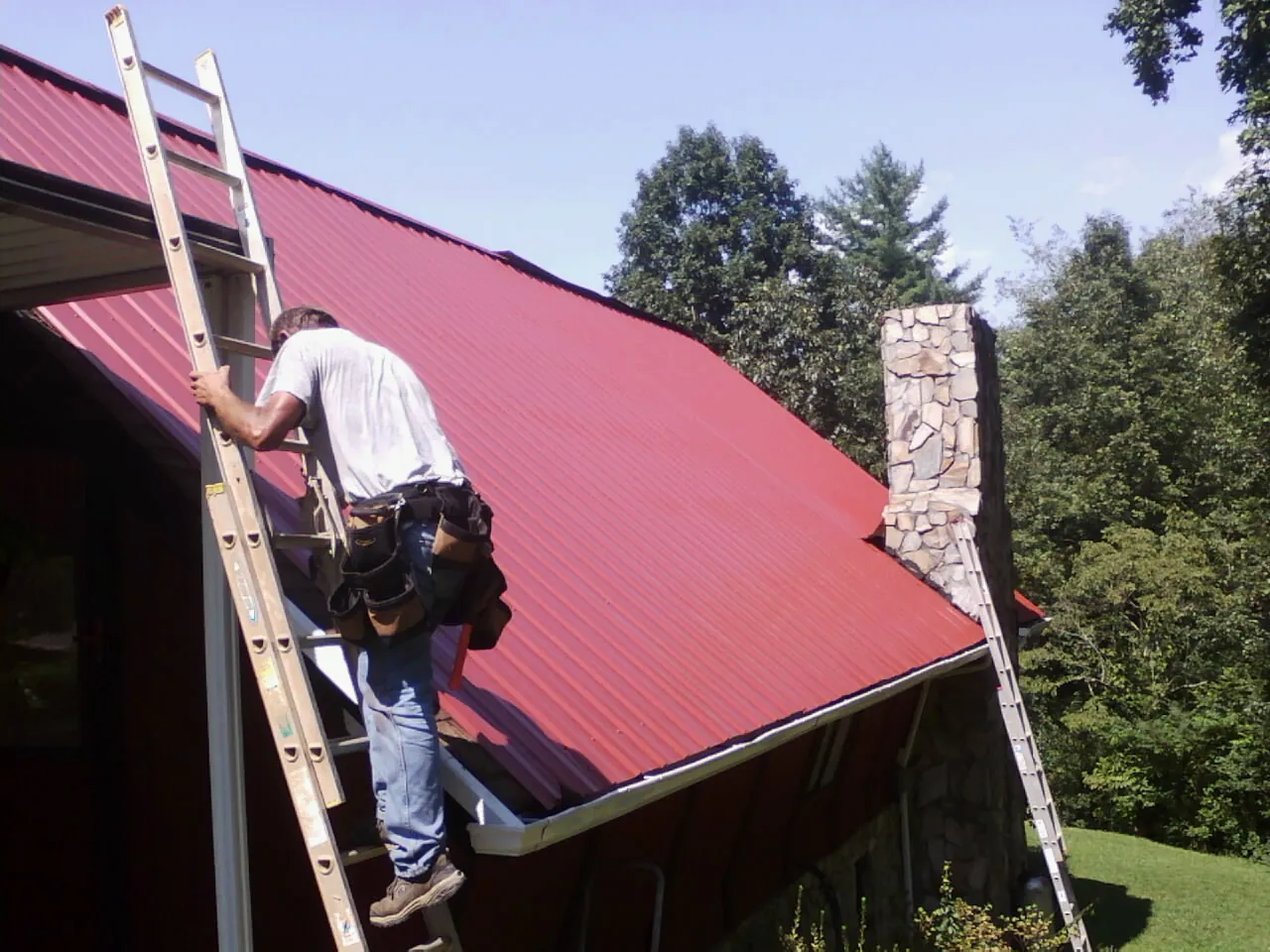 Roofer on a ladder beside a red metal roof and stone chimney — Soffit & Fascia Repair by Roofing Brothers in Radnor